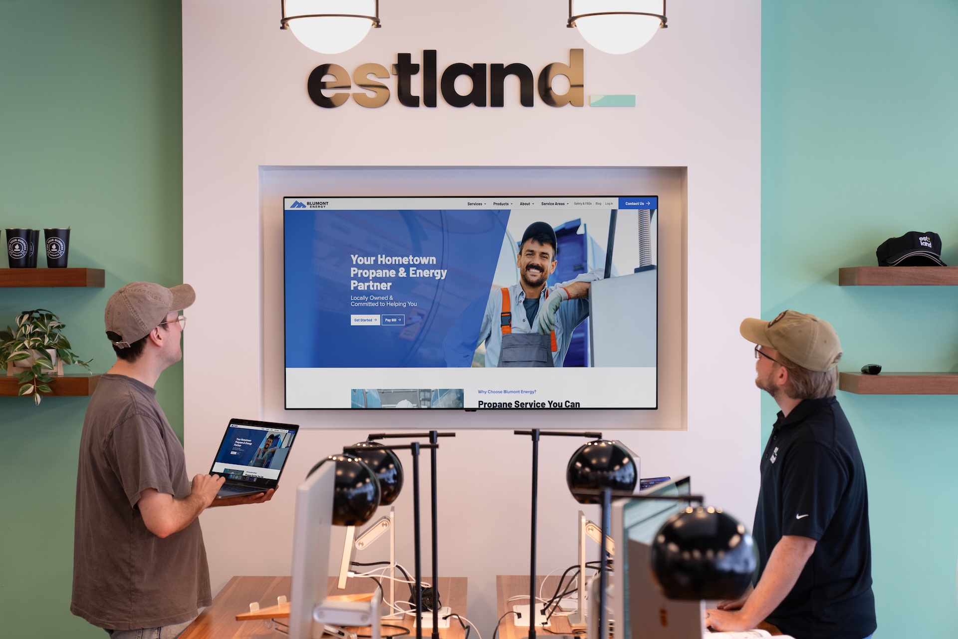 Two men are in Estland's office, focused on a large screen displaying a website development mockup. One man holds a laptop while the other looks at the screen.