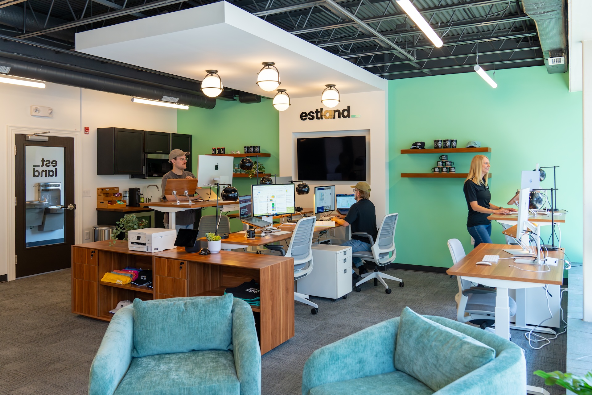 Estland's new office space features modern desks, computers and shelves. Two employees are working at their desks while another stands nearby at a workstation. The walls are painted in a light green color, and there are comfortable chairs in the foreground.