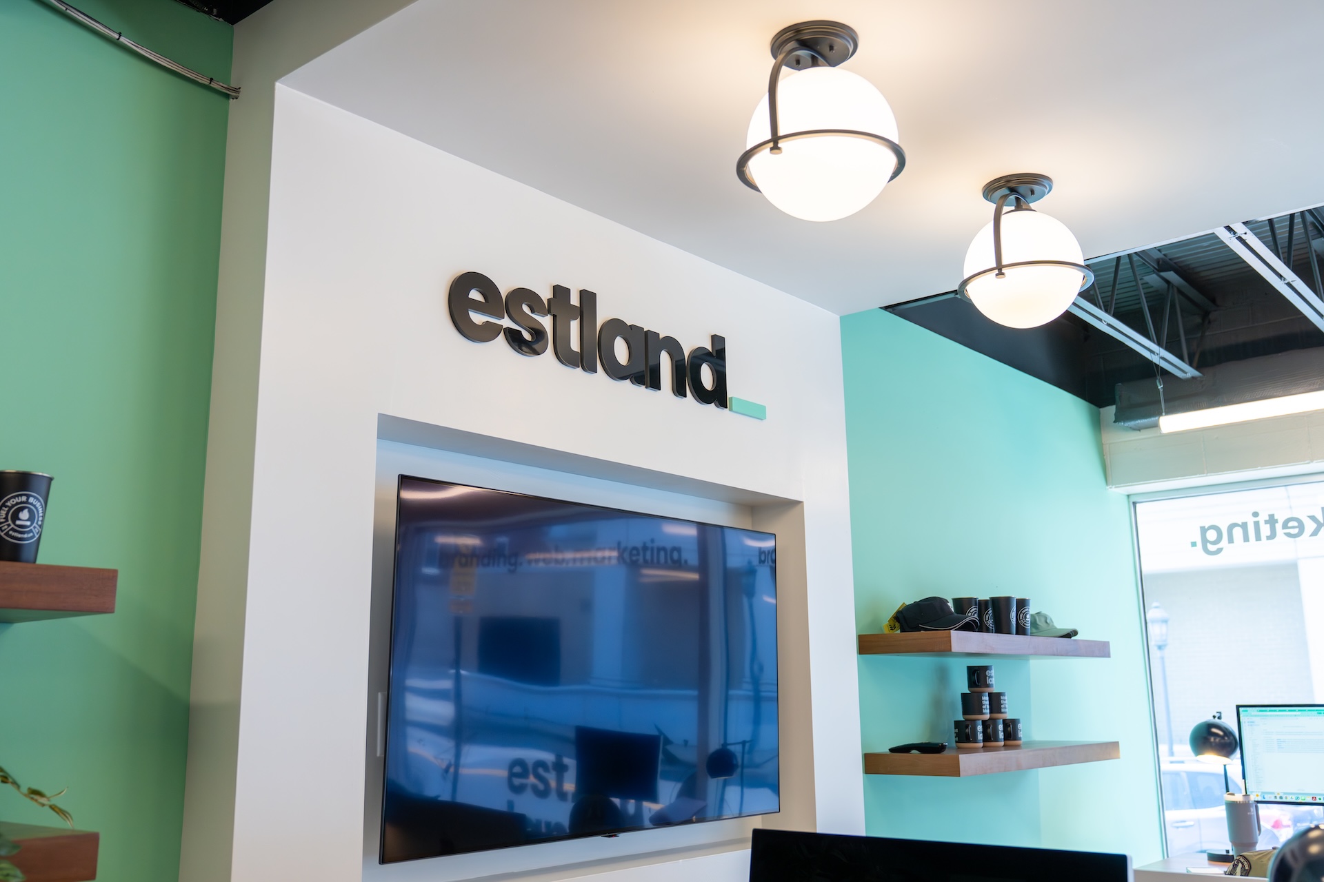 Estland's new marketing office features a wall with the text "estland" in a stylized font. Below the text, there is a flat-screen TV mounted on a white wall. The background features green walls and decorative shelves displaying various items, while light fixtures hang from the ceiling.