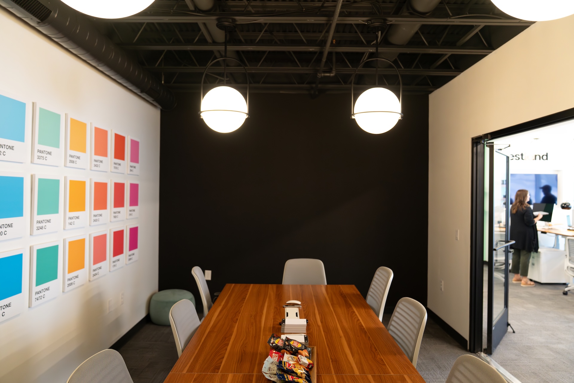 Estland's conference room featuring a large wooden table surrounded by several chairs. On one wall, a series of colorful rectangular panels is displayed. The opposite wall is painted black, and two round overhead lights provide illumination.
