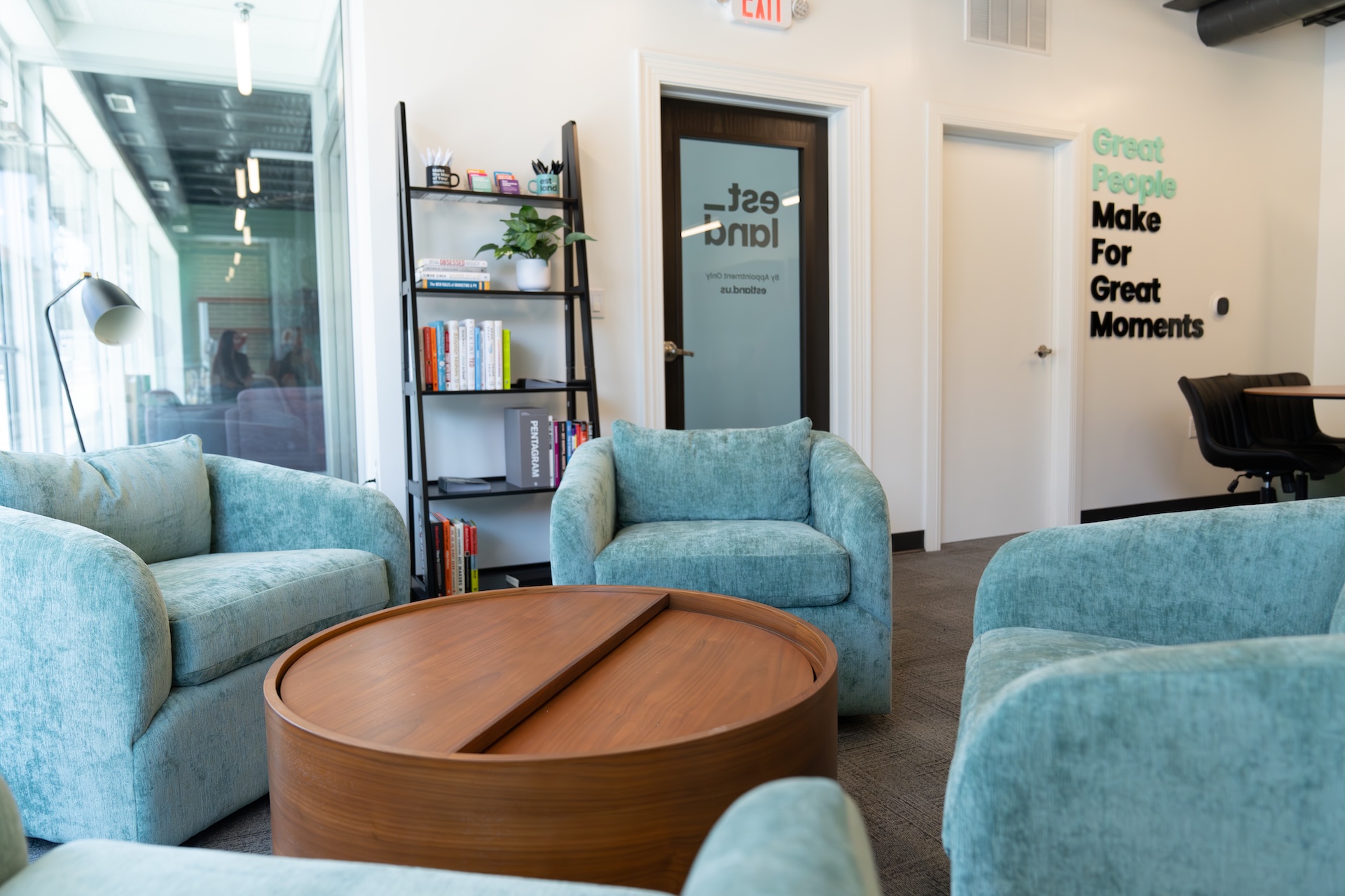 Estland's office lounge featuring light green upholstered chairs arranged around a wooden coffee table. A bookshelf in the background holds various marketing books.