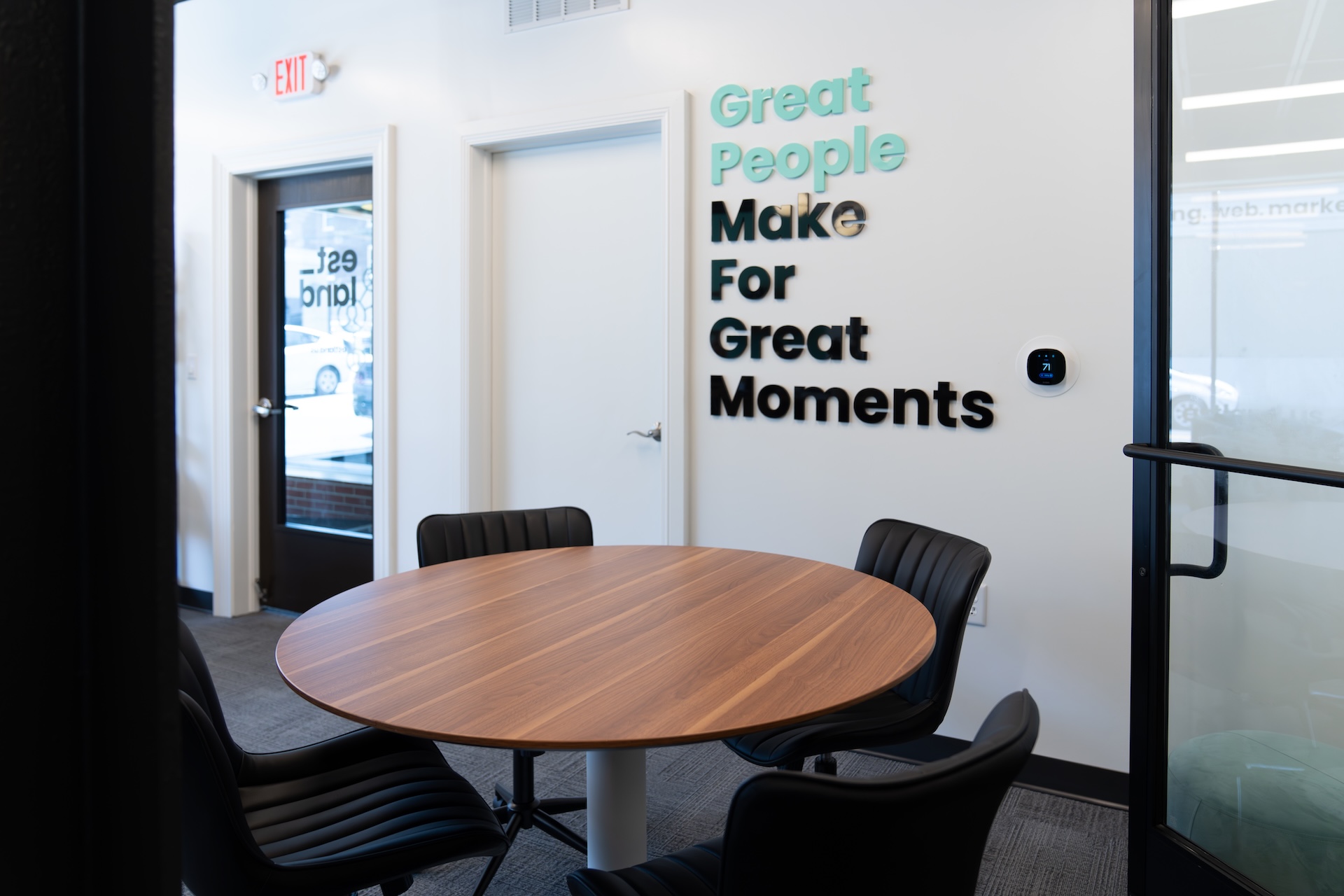 Estland's office features a round wooden table surrounded by black chairs. A wall displays the phrase "Great People Make For Great Moments" in stylized lettering.