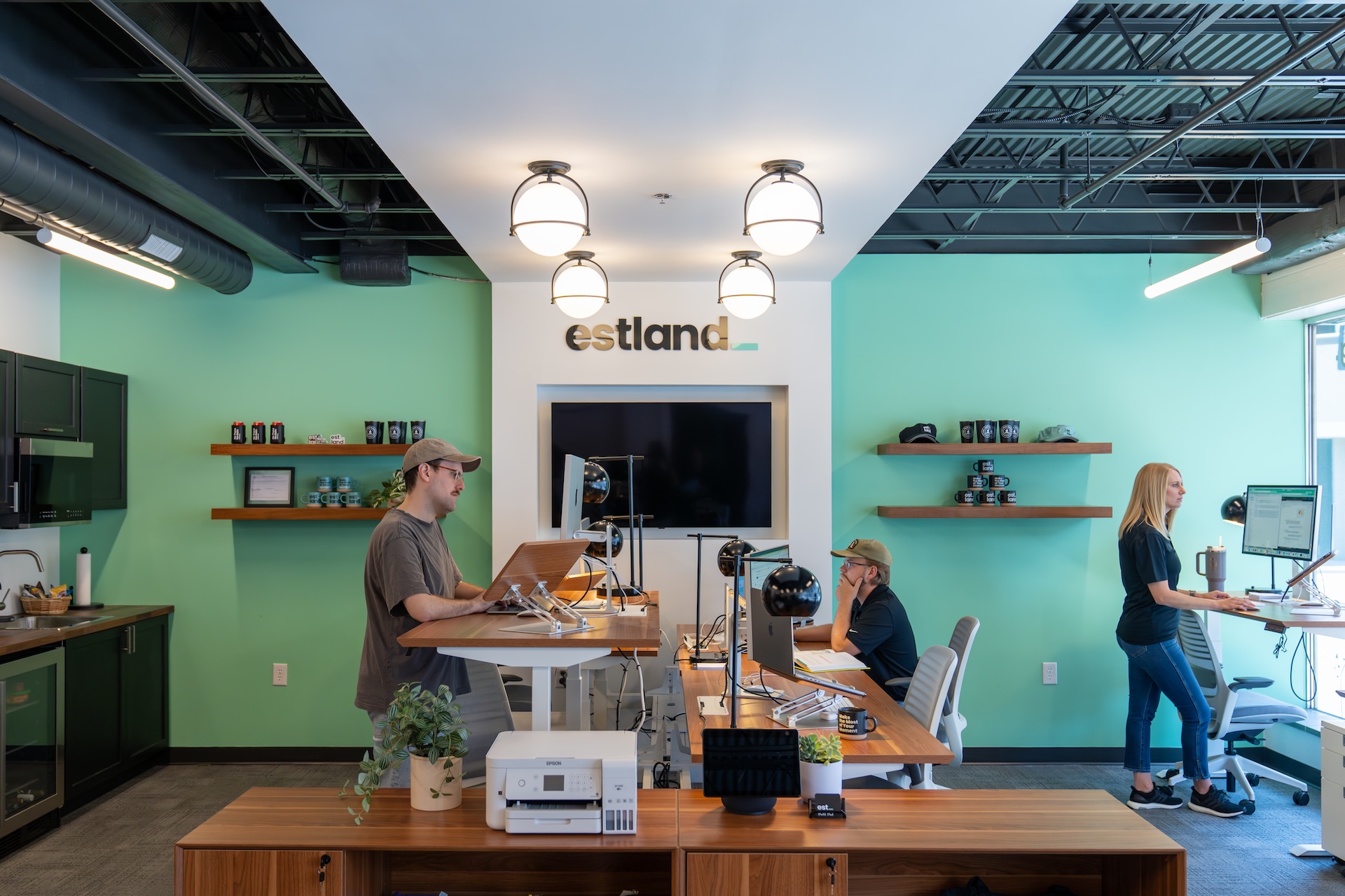 Estland's new office space features a green accent wall. In the foreground, people are working at marketing and branding work stations. Shelves display various items, and a large screen is mounted on the wall above.