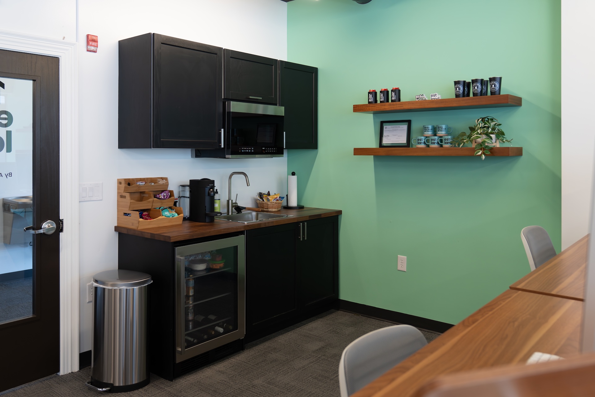 A modern kitchen area featuring black cabinets and a wooden countertop. The wall is painted green. A sink is integrated into the cabinetry, which also includes a microwave and a small refrigerator. Shelves above the counter display several jars and decorative items.