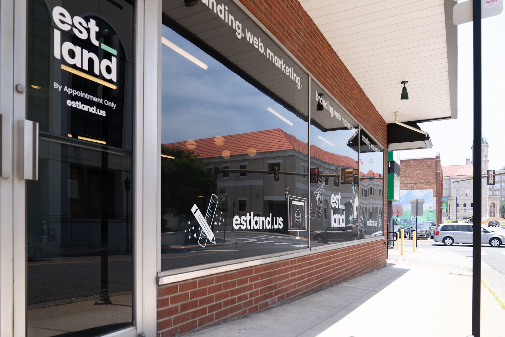 A storefront with large glass windows displaying the logo and signage of "estland," a web marketing company. The reflections in the windows show a city street with buildings in the background and parked cars. The setting is bright and appears to be a sunny day.