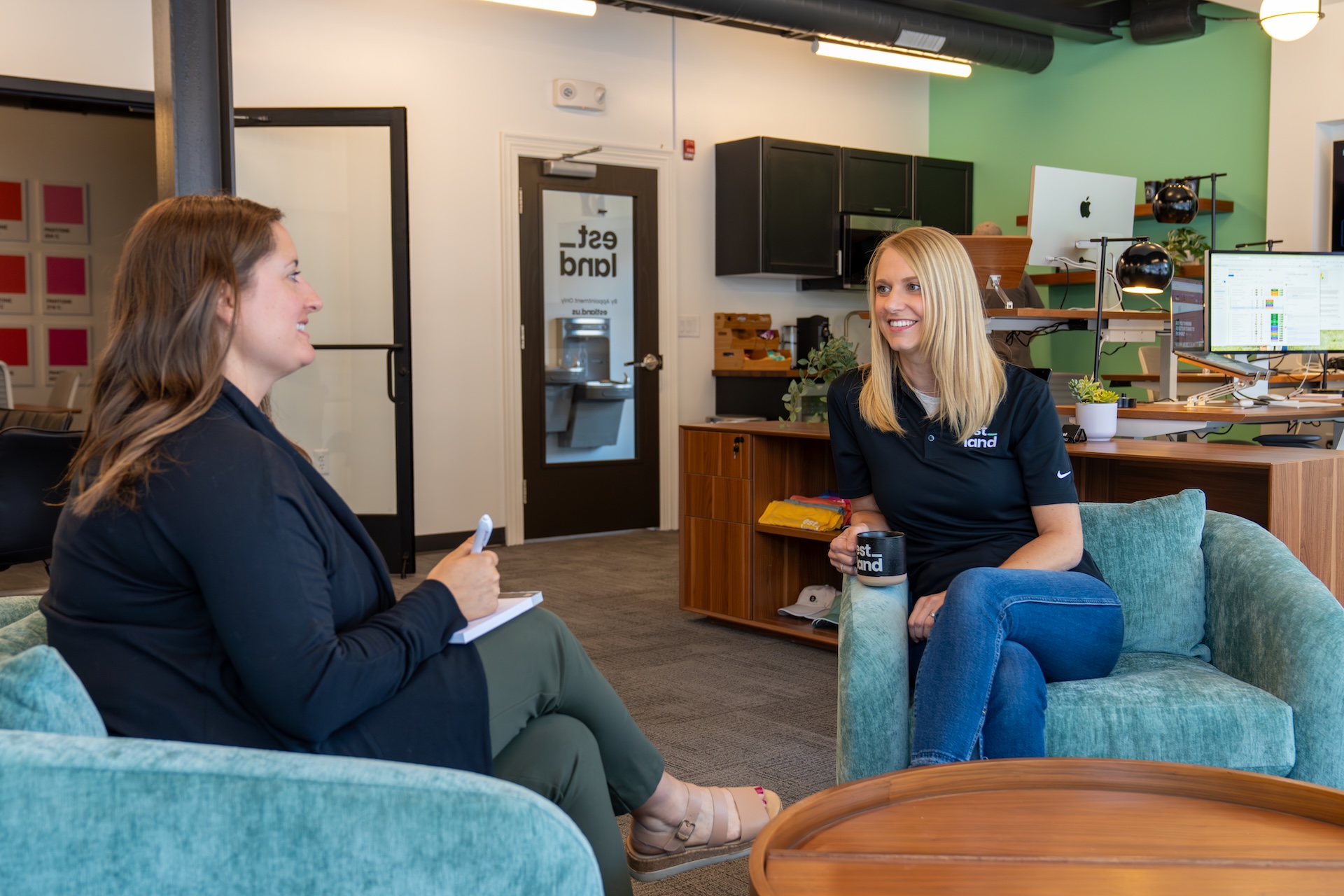 Two women sit in Estland's new office space. One is taking notes while the other smiles and engages in conversation. The background features modern furniture and a large window, creating a professional yet inviting atmosphere.